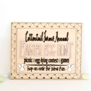 Puede incluir: Un letrero de madera con un borde tejido, anunciando una b&uacute;squeda anual de huevos de Pascua de Cottontail Farms. El letrero presenta un dise&ntilde;o de huevo de Pascua rosa y negro, siluetas de conejos y el texto "picnic egg dying contest games hop on over for some fun".