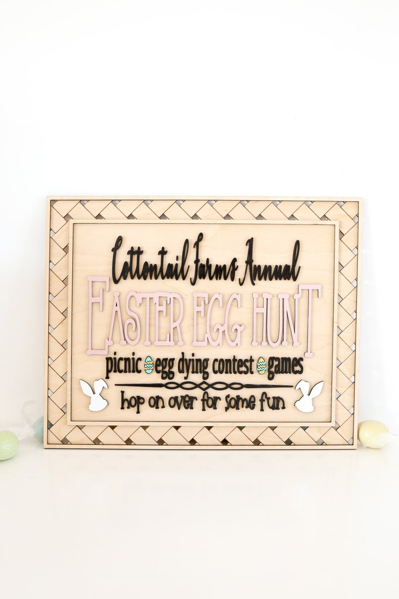 Puede incluir: Un letrero de madera con un borde tejido, anunciando una b&uacute;squeda anual de huevos de Pascua de Cottontail Farms. El letrero presenta un dise&ntilde;o de huevo de Pascua rosa y negro, siluetas de conejos y el texto "picnic egg dying contest games hop on over for some fun".