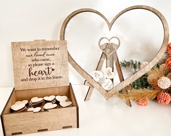 May include: A wooden box with a sign that reads "We want to remember our loved ones who came, so please sign a heart and drop it in this frame." The box is filled with small wooden hearts. A wooden heart-shaped frame with a glass insert is on a stand. The frame is filled with signed wooden hearts.