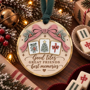 May include: A round wooden Christmas ornament with a gold metal rim and a dark green velvet ribbon. The ornament features a pink bow, three mahjong tiles with holiday designs, and the text "Good tiles GREAT FRIENDS best memories".