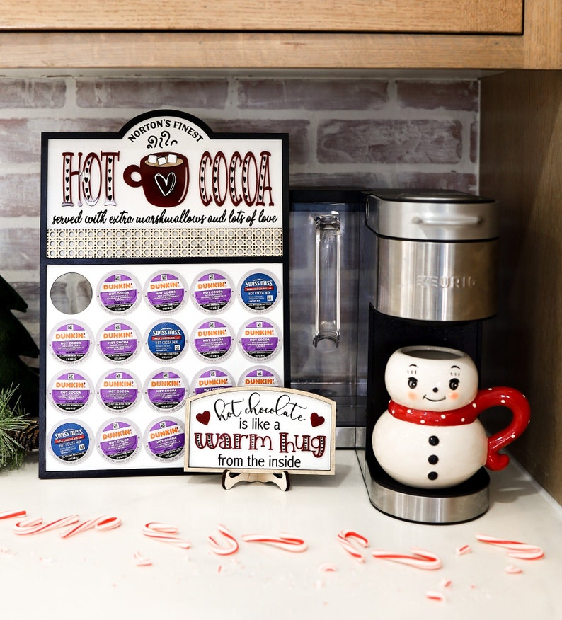 May include: A black sign with white lettering that reads "Norton's Finest Hot Cocoa served with extra marshmallows and lots of love". The sign has a picture of a mug with a heart on it. There are 24 coffee pods in a grid on the sign. A white sign with red hearts and black lettering that reads "Hot chocolate is like a warm hug from the inside" is in front of a silver coffee maker with a snowman mug on top. There are broken candy canes on the counter.