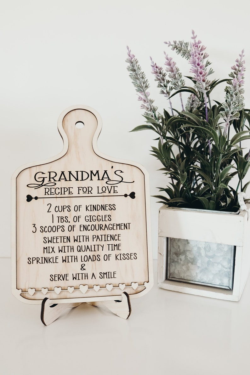 May include: A wooden sign shaped like a cutting board with the text "Grandma's Recipe for Love" and a list of ingredients for love, including kindness, giggles, encouragement, patience, quality time, and kisses. The sign is on a small wooden stand and has a row of small hearts around the bottom edge.