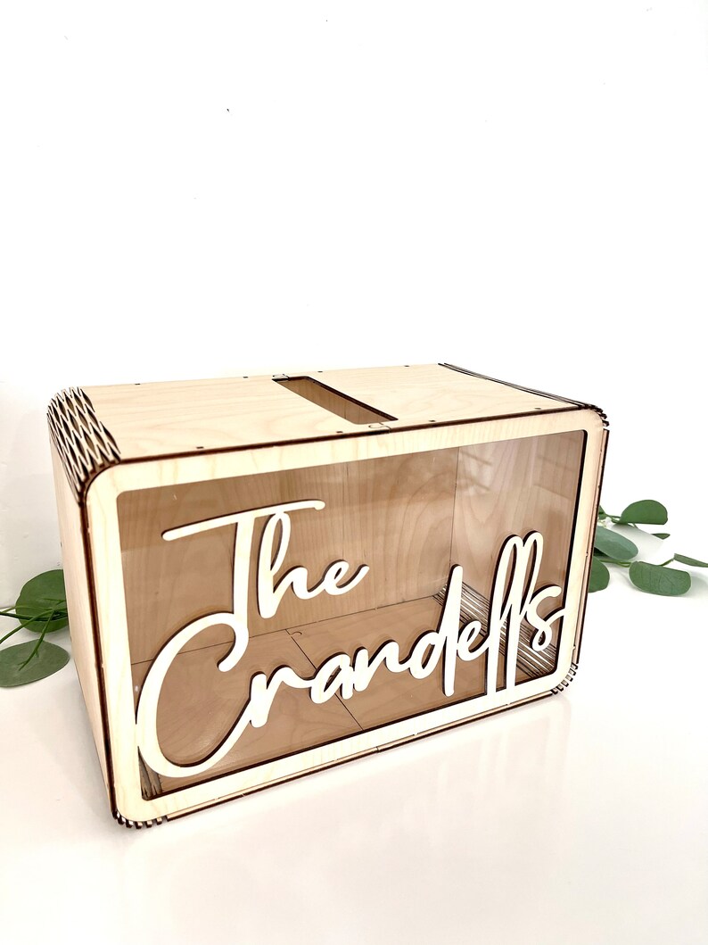 May include: A wooden box with a clear acrylic front panel. The box has a slot on the top for inserting cards or envelopes. The text "The Crandells" is cut out of the front panel.