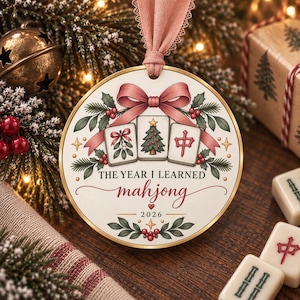 May include: A round white Christmas ornament with a gold rim and a pink ribbon hanger. The design features three mahjong tiles, a pink bow, holly leaves, and berries. Text reads: THE YEAR I LEARNED mahjong 2026, with a small red heart above the year.