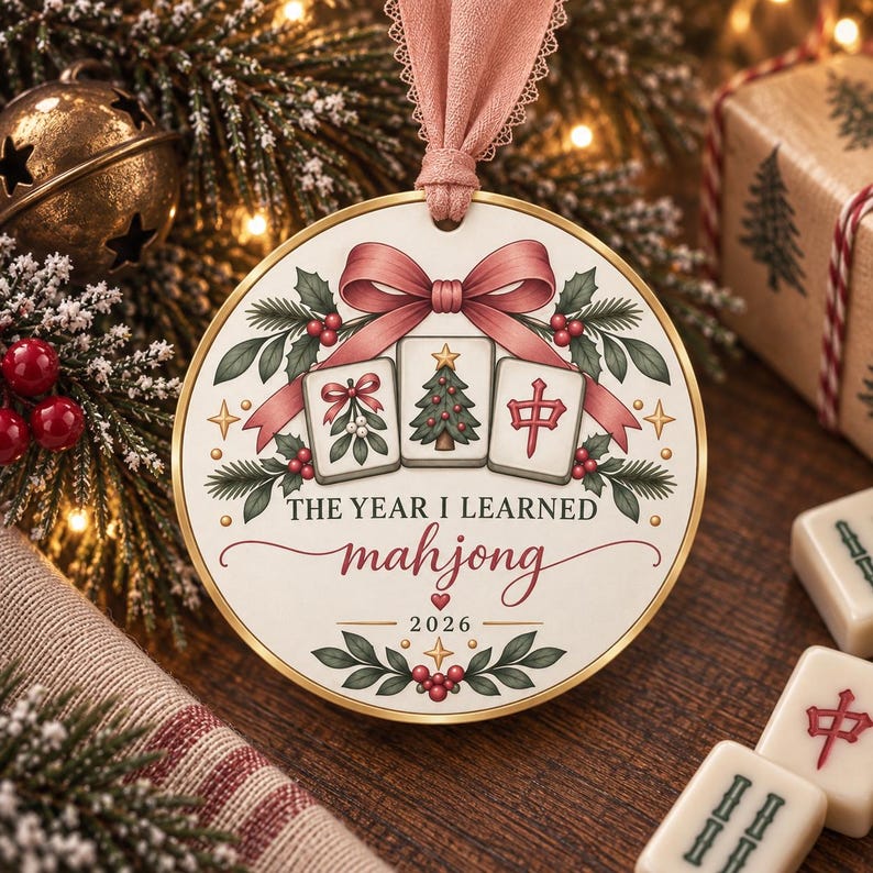 May include: A round white Christmas ornament with a gold rim and a pink ribbon hanger. The design features three mahjong tiles, a pink bow, holly leaves, and berries. Text reads: THE YEAR I LEARNED mahjong 2026, with a small red heart above the year.