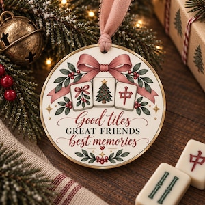 May include: A round, cream-colored Christmas ornament featuring a pink ribbon hanger. The design includes three mahjong tiles, holly leaves, and berries, with the text "Good tiles GREAT FRIENDS best memories" printed in dark red and black script.