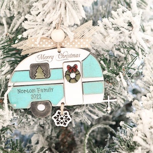 May include: A teal and white wooden Christmas ornament shaped like a vintage camper with the text "Merry Christmas" and "Norton Family 2022". The ornament has a red bow and a green wreath on the door. A snowflake charm hangs from the bottom.