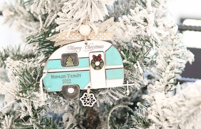 May include: A teal and white wooden Christmas ornament shaped like a vintage camper with the text "Merry Christmas" and "Norton Family 2022". The ornament has a red bow and a green wreath on the door. A snowflake charm hangs from the bottom.