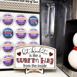 May include: A wooden sign with a white background and red hearts reads "hot chocolate is like a warm hug from the inside". The sign is on a stand and in front of a coffee maker and a display of coffee pods.