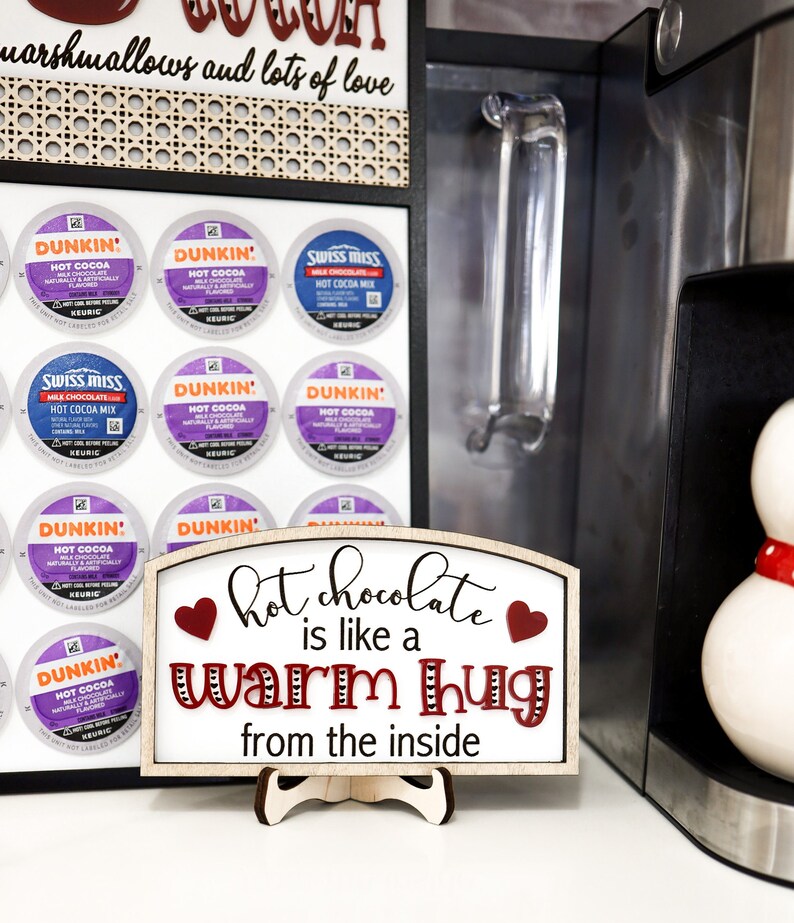 May include: A wooden sign with a white background and red hearts reads "hot chocolate is like a warm hug from the inside". The sign is on a stand and in front of a coffee maker and a display of coffee pods.