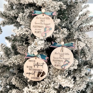May include: Two wooden Christmas ornaments with a blue and pink ribbon. The ornaments are round and have a white background with black text. The text on the ornaments reads: "The tiniest little feet leave the biggest footprints on our hearts" and "There is no footprint too small that it cannot leave an imprint on the world".