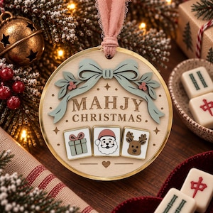 May include: A round wooden Christmas ornament with a gold rim and a pink ribbon hanger. The ornament features a light green bow, the text "MAHJY CHRISTMAS," and three tiles depicting a gift, a Santa Claus face, and a reindeer face.