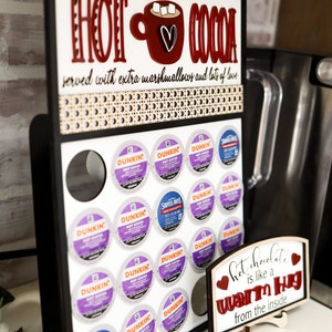 May include: A black wooden sign with the text "Norton's Finest Hot Cocoa" and a red mug with marshmallows and a heart. The sign has a white background and is decorated with a brown and white patterned border. The sign is mounted on a white surface and has a display of coffee pods underneath. The coffee pods are in a variety of colors and brands, including Dunkin' and Swiss Miss. A small wooden sign with the text "Hot chocolate is like a warm hug from the inside" is in the foreground.