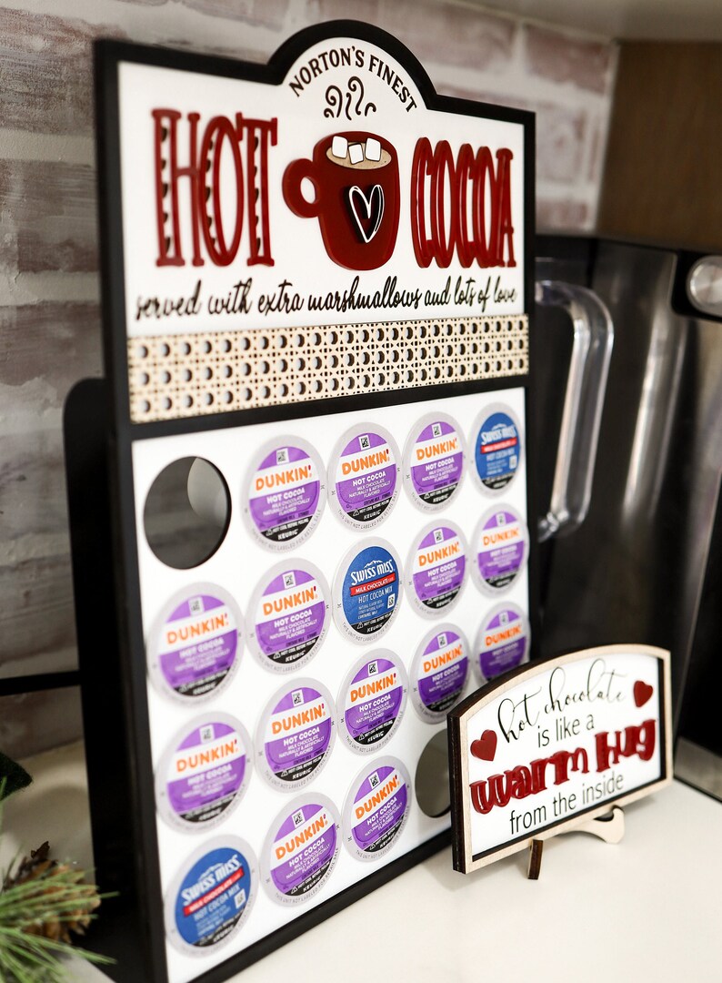 May include: A black wooden sign with the text "Norton's Finest Hot Cocoa" and a red mug with marshmallows and a heart. The sign has a white background and is decorated with a brown and white patterned border. The sign is mounted on a white surface and has a display of coffee pods underneath. The coffee pods are in a variety of colors and brands, including Dunkin' and Swiss Miss. A small wooden sign with the text "Hot chocolate is like a warm hug from the inside" is in the foreground.