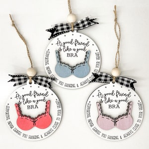 May include: Three white round ornaments with black and white checkered ribbon. Each ornament has a different colored bra cutout with the text "A good friend is like a good BRA" and "SUPPORTIVE NEVER LEAVES YOU HANGING & ALWAYS CLOSE TO YOUR HEART".