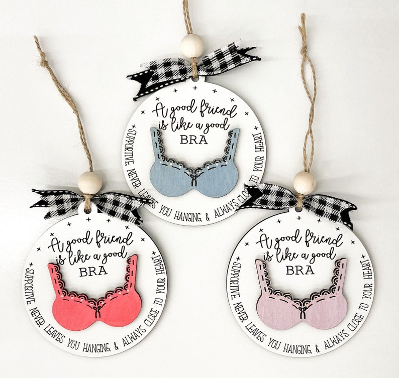 May include: Three white round ornaments with black and white checkered ribbon. Each ornament has a different colored bra cutout with the text "A good friend is like a good BRA" and "SUPPORTIVE NEVER LEAVES YOU HANGING & ALWAYS CLOSE TO YOUR HEART".