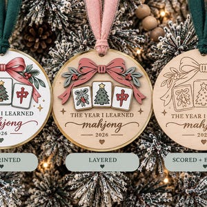 May include: Three round wooden ornaments with green or pink ribbons. Each features a bow, holly, and three mahjong tiles with a Christmas tree and the text THE YEAR I LEARNED mahjong 2026. Styles include UV printed, layered, and scored and engraved.