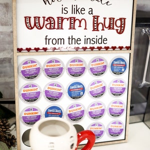 May include: A wooden sign with the text "hot chocolate is like a warm hug from the inside" and a snowman mug. The sign has a shelf with 15 single-serve coffee pods, including Dunkin' and Swiss Miss hot chocolate.