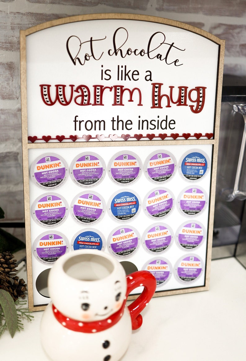 May include: A wooden sign with the text "hot chocolate is like a warm hug from the inside" and a snowman mug. The sign has a shelf with 15 single-serve coffee pods, including Dunkin' and Swiss Miss hot chocolate.