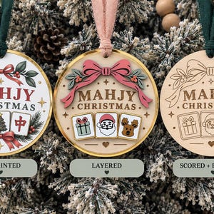 May include: Three round ornaments labeled UV PRINTED, LAYERED, and SCORED + ENGRAVED. Each features the text MAHJY CHRISTMAS, a bow, and three mahjong tiles. The first is printed, the second has layered elements, and the third is laser engraved.