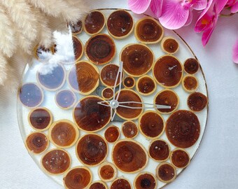 May include: A circular wall clock featuring numerous small, round wood slices embedded in a clear, glossy resin base. The clock has silver-toned metal hands positioned in the center, displaying a natural wood grain pattern throughout the design.