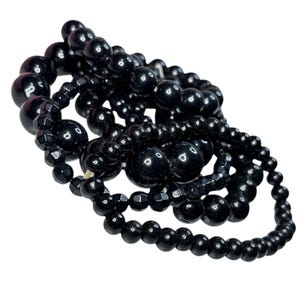 May include: A selection of black beaded necklaces and bracelets. The beads are of various sizes and shapes, including round and faceted designs. The jewellery is arranged in a layered, intertwined manner, creating a visually rich display.