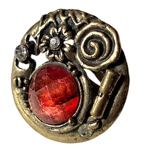 May include: A circular brass-toned metal button featuring a large, faceted red stone at the centre. The surface is decorated with raised floral and spiral patterns, accented by three small, clear embedded crystals along the outer edge.