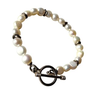 Puede incluir: Pulsera de perlas blancas irregulares con detalles plateados y cierre de palanca. Las perlas, de forma ovalada, son de un blanco nacarado. El cierre es redondo y plateado. Una joya elegante y atemporal.