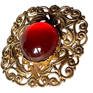 Puede incluir: Un broche de tono dorado que presenta un marco de filigrana calado e intrincado con motivos florales y de volutas. Una piedra cabujón grande, lisa, redonda y de color rojo intenso está engastada prominentemente en el centro.