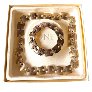 May include: A gold-toned jewelry set, including a necklace and bracelet, presented in a cream-colored box. The necklace and bracelet feature square-shaped links with embedded clear crystals. The set is displayed on a white background.