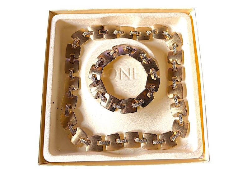 May include: A gold-toned jewelry set, including a necklace and bracelet, presented in a cream-colored box. The necklace and bracelet feature square-shaped links with embedded clear crystals. The set is displayed on a white background.
