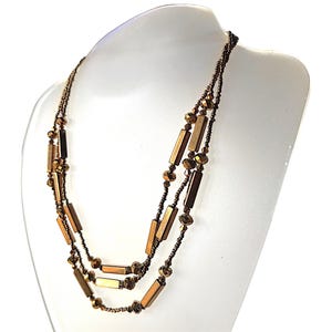 May include: A multi-strand necklace featuring metallic bronze-toned rectangular bar beads and smaller faceted round beads. The design consists of three layered strands of varying lengths, showcasing a consistent warm copper colour palette throughout.