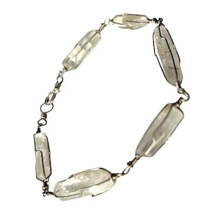 Può includere: Un braccialetto argentato con perline di cristallo oblunghe e trasparenti, racchiuse in filo metallico. I cristalli sono collegati da piccole catene e chiusure argentate, creando un accessorio delicato ed elegante. Il braccialetto è su sfondo bianco.