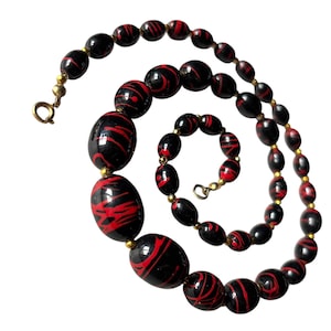 May include: A black beaded necklace and bracelet set with red swirl patterns. The necklace and bracelet have gold-colored clasps and connectors. The beads are oval and round, with a glossy finish.