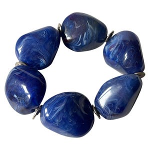 Op de afbeelding: Een opvallende armband met zes grote, ovale kralen in een diepblauwe kleur met wervelende witte patronen. De kralen zijn verbonden door kleine, zilverkleurige metalen ringen, wat een opvallend accessoire creëert.