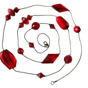 May include: A long necklace featuring a thin silver-toned chain interspersed with various red translucent beads. The beads include faceted geometric shapes, smooth round spheres, and elongated oval forms. A metal lobster claw clasp is attached.