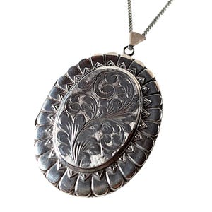 May include: Oval silver locket pendant with intricate floral engraving. The locket hangs from a delicate silver chain. The edges of the locket are scalloped, adding a decorative touch. The pendant is a classic piece of jewelry.