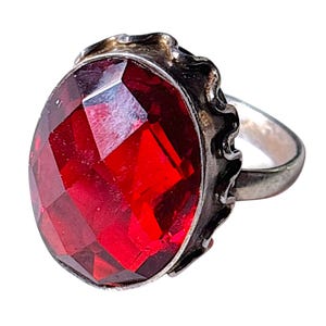 May include: A silver-toned ring featuring a large, oval-shaped, faceted red gemstone set in a scalloped metal bezel. The band is plain and metallic, supporting the prominent, deep red stone which displays multiple geometric facets across its surface.