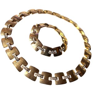 May include: A gold-toned necklace and bracelet set. The jewelry features a series of square-shaped links with a brushed finish, each accented with small, sparkling stones. The necklace is curved, and the bracelet is circular.