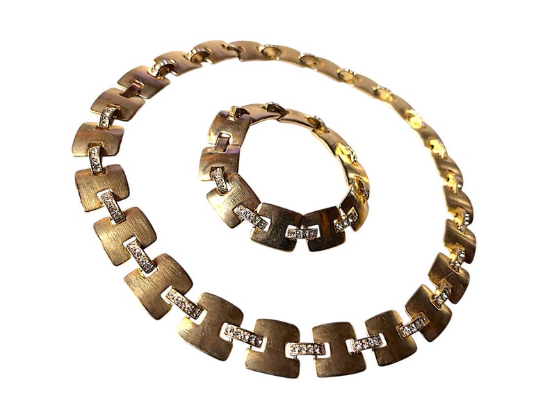 May include: A gold-toned necklace and bracelet set. The jewelry features a series of square-shaped links with a brushed finish, each accented with small, sparkling stones. The necklace is curved, and the bracelet is circular.