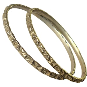 May include: Two gold-toned metal bangles featuring a repeating geometric pattern of etched triangles and lines along the outer surface. The bracelets have a smooth, polished interior and a textured, decorative exterior with a metallic finish.