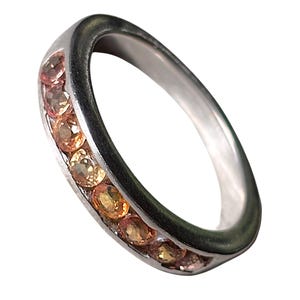 May include: A silver-toned metal ring featuring a channel setting with a row of seven round-cut, warm-toned orange and peach gemstones. The band has a smooth, polished finish and a rounded profile, showcasing the stones embedded along the outer curve.