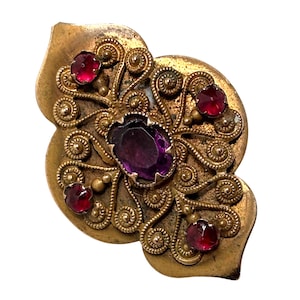 Puede incluir: Broche dorado ornamentado con un diseño floral. El broche presenta una gran gema púrpura ovalada en el centro, rodeada de gemas rojas más pequeñas y un intrincado trabajo de volutas. Un accesorio de inspiración vintage.