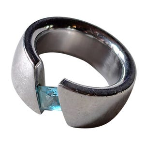 May include: A silver-toned metal ring with a wide, open-ended band design. The ring features a single, square-cut light blue gemstone set between the two ends of the band, creating a tension-style setting with a smooth, polished finish.
