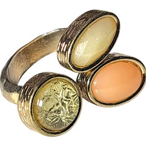 May include: A silver-toned metal ring featuring an open-ended design with three distinct settings. One setting is a round, textured yellow stone, while the other two are smooth, oval-shaped cabochons in pale yellow and soft peach colours.
