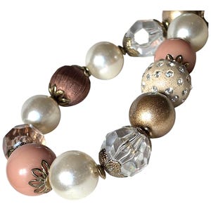 Könnte beinhalten: Ein Armband mit Perlen in Weiß, Rosa, Braun und Gold. Die Perlen umfassen perlmuttartige, facettierte und strukturierte Designs. Einige Perlen sind mit Strasssteinen und Metalldetails verziert. Dieses Armband ist für verschiedene Anlässe geeignet.