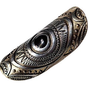 May include: A long, curved silver-toned metal ring featuring an elongated oval shape. The surface is decorated with intricate, raised geometric patterns, including small dots and lines, and centres on a single, smooth, dark black circular stone.