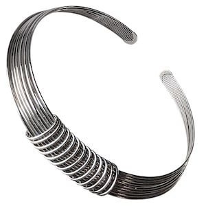 May include: A silver-toned metal cuff bracelet featuring a multi-strand design. The bracelet consists of several thin, parallel wires that are gathered and secured in the centre by a tightly coiled silver wire wrap, creating a layered appearance.