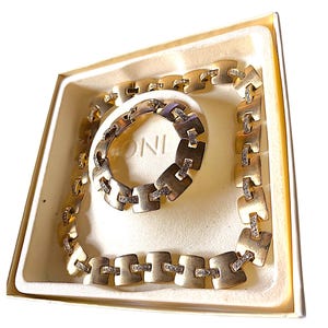 May include: A gold-toned necklace and bracelet set with square links and small, clear stones. The jewelry is displayed in a cream-colored box with the word "ONI" visible on the bracelet.