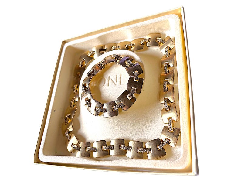 May include: A gold-toned necklace and bracelet set with square links and small, clear stones. The jewelry is displayed in a cream-colored box with the word "ONI" visible on the bracelet.
